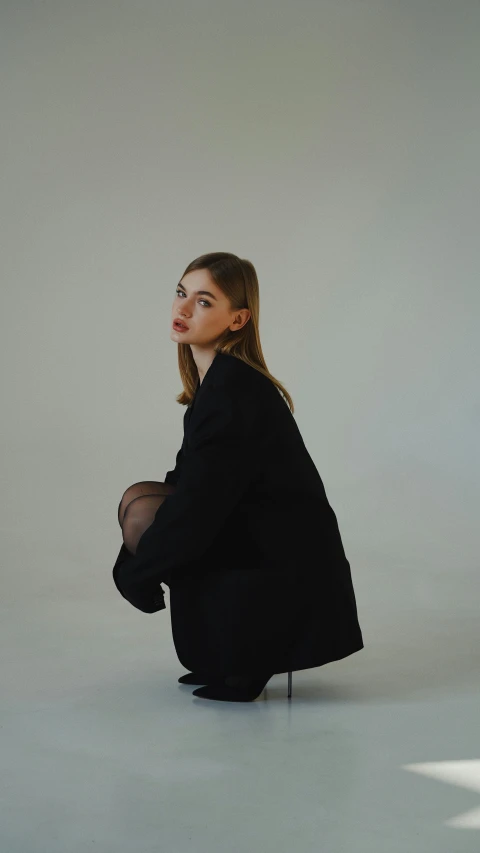 a woman kneeling down in a white room, an album cover, unsplash, wearing black suit, chloë grace moretz, short skirt and a long jacket, press shot