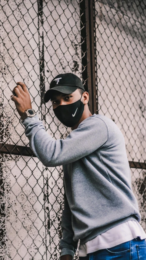 a man wearing a face mask leaning against a fence, an album cover, by Robbie Trevino, trending on pexels, nike logo, casual black clothing, discord profile picture, gray canvas