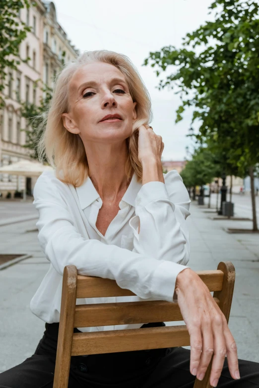 a woman sitting on top of a wooden bench, a portrait, by Matija Jama, trending on unsplash, white hair color, wearing a white blouse, in a city square, sitting on designer chair