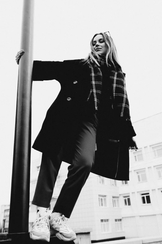 a black and white photo of a woman leaning on a pole, inspired by Peter Lindbergh, unsplash, burberry, flare, wearing black coat, a blond