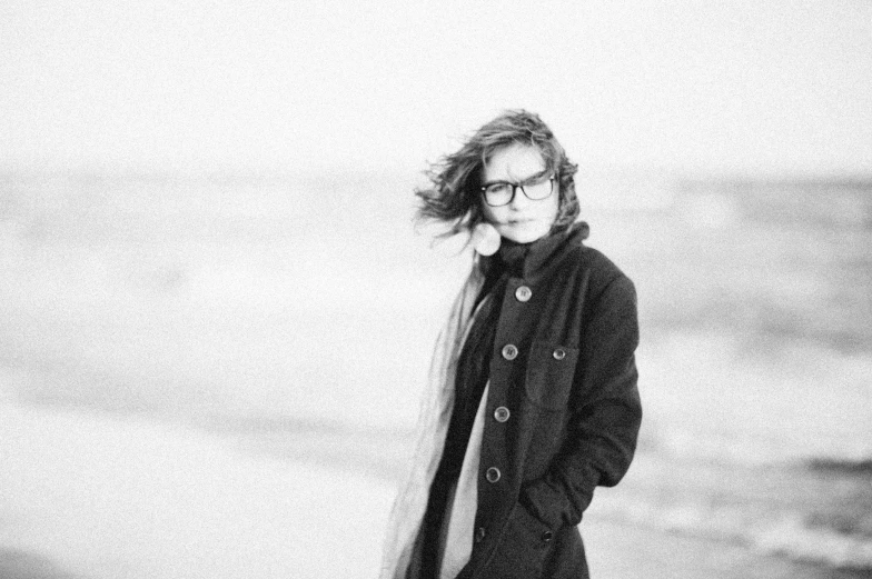 a woman standing on top of a beach next to the ocean, a black and white photo, by Karl Buesgen, pexels, girl with glasses, wearing black overcoat, wind - swept, portrait androgynous girl