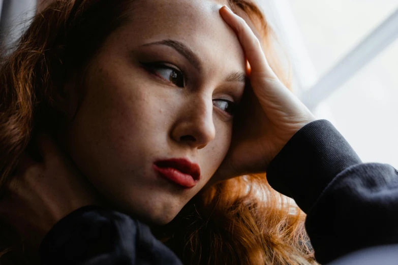 a woman sitting in front of a window with her hand on her head, trending on pexels, ( redhead, exhausted face close up, extremely graphic, thumbnail