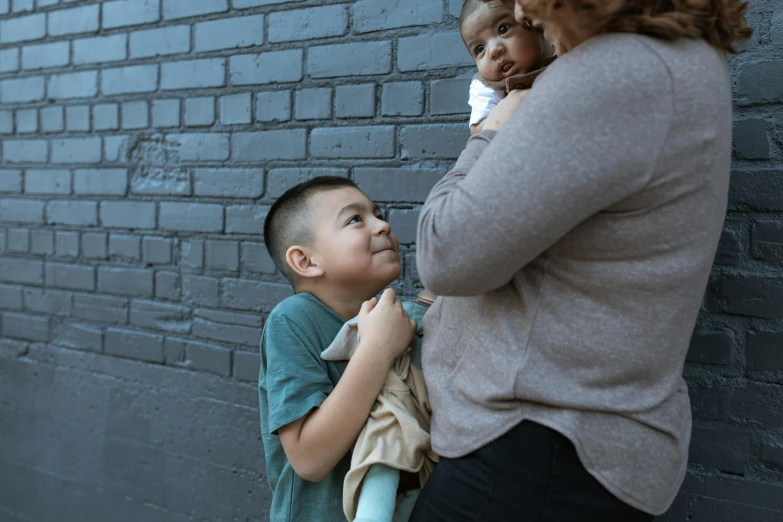 a woman standing next to a boy in front of a brick wall, by Winona Nelson, pexels contest winner, incoherents, an obese, hugs, towering above a small person, varying ethnicities