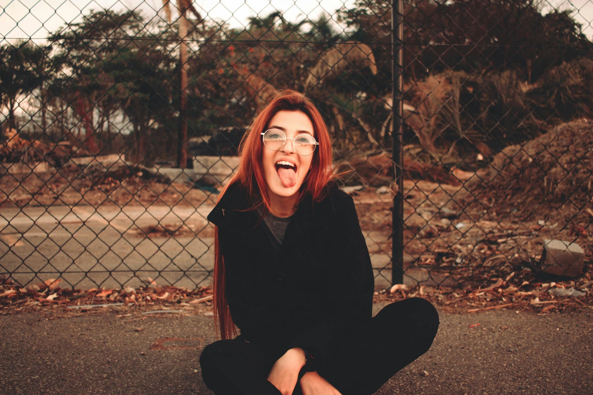 a woman sitting on the ground in front of a fence, trending on pexels, antipodeans, tongue out, ( redhead, happy trippy mood, wearing black glasses