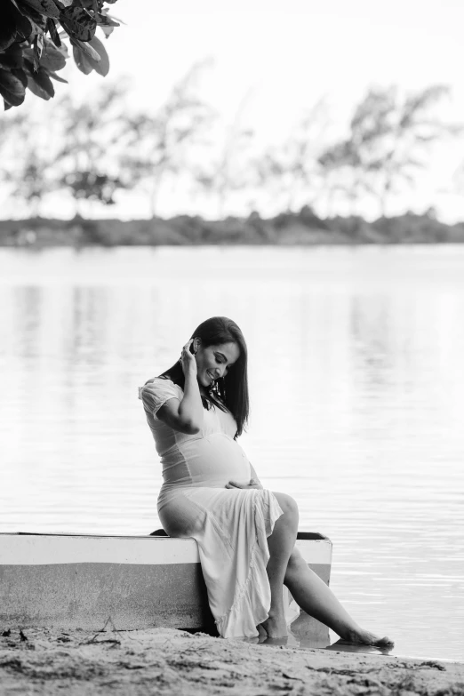 a pregnant woman sitting on a bench next to a body of water, a black and white photo, unsplash, various poses shooting photos, maternal, belle, album cover