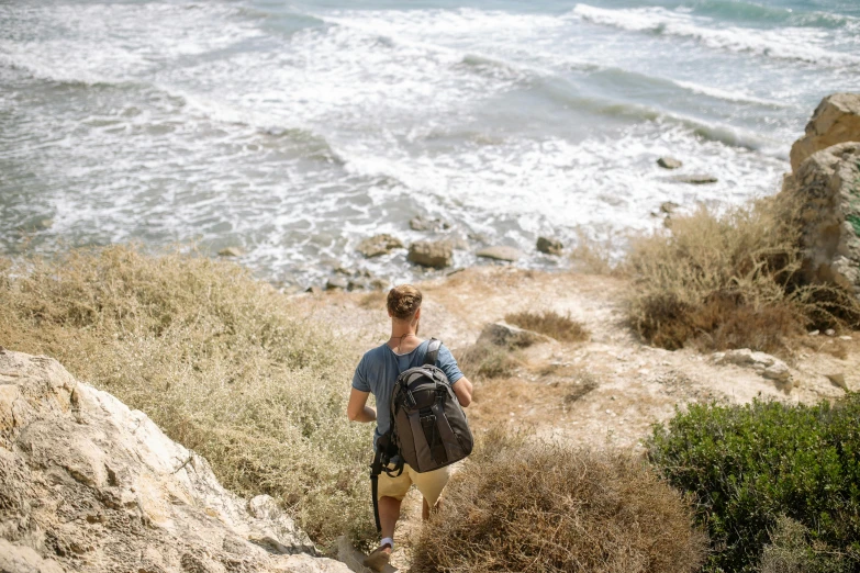 a man walking up a steep hill next to the ocean, pexels contest winner, happening, with a backpack, cyprus, avatar image, epic coves crashing waves plants
