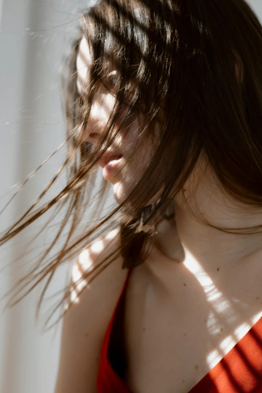 a woman in a red dress standing in front of a window, trending on pexels, visual art, hands in her hair. side-view, in a sunbeam, portrait sophie mudd, the wind moves her shirt