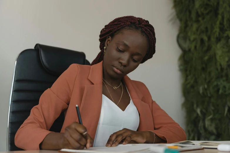 a woman sitting at a desk writing on a piece of paper, by Chinwe Chukwuogo-Roy, pexels contest winner, girl in suit, brown skin, official screenshot, colored