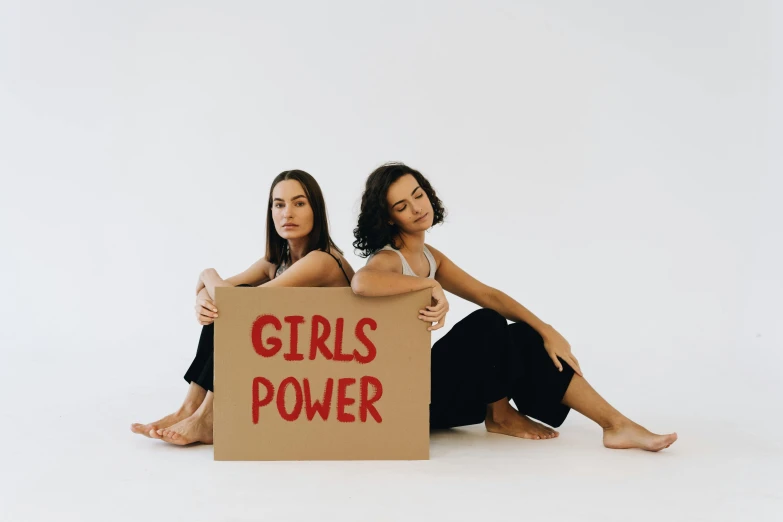 two women sitting next to each other holding a sign, trending on pexels, feminist art, power pose, background image, girl standing, bella poarch