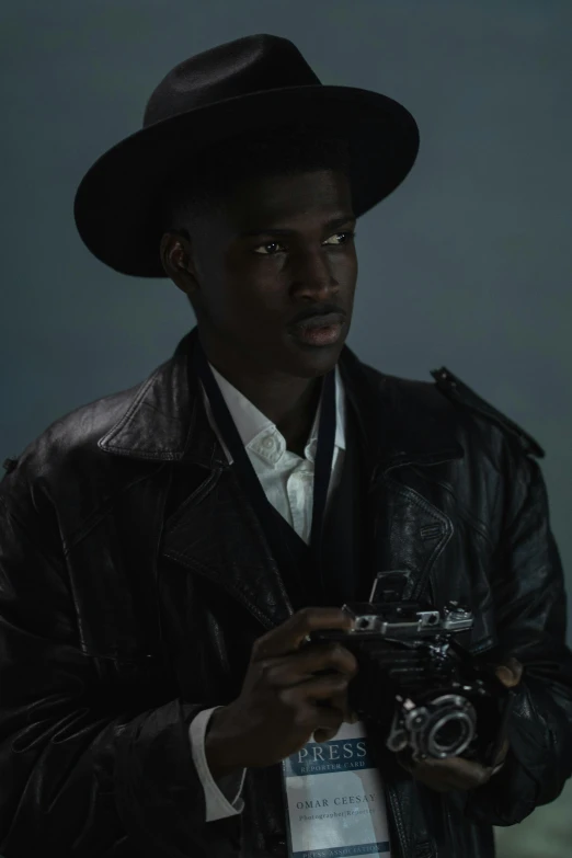 a man in a hat holding a camera, inspired by Gordon Parks, afrofuturism, adut akech, wearing a leather trench coat, cowboy shot, ( ( theatrical ) )