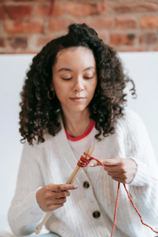 a woman knitting in front of a brick wall, a portrait, trending on pexels, floggers, wearing a white sweater, avatar image, mixed-race woman