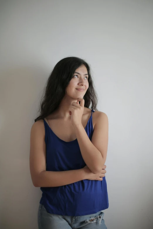 a woman standing in front of a white wall, pointed chin, female with long black hair, thoughtful, wearing blue dress