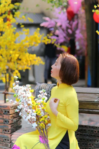 a woman sitting on a bench holding a bunch of flowers, inspired by Cui Bai, pexels contest winner, happening, yellow clothes, ao dai, sakura tree in background, vivid)