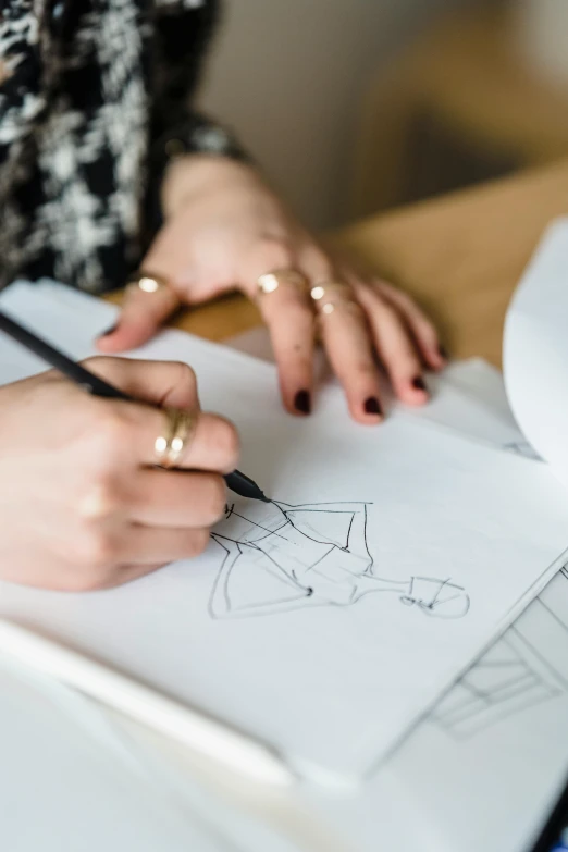 a woman sitting at a table drawing on a piece of paper, a drawing, by Nicolette Macnamara, trending on pexels, tailored clothing, fancy line work, brittney lee, costume desig