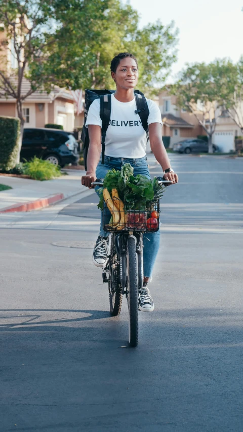 a woman riding a bike down a street, by Josh Bayer, pexels contest winner, confident holding vegetables, graphic tees, los angeles ca, official screenshot