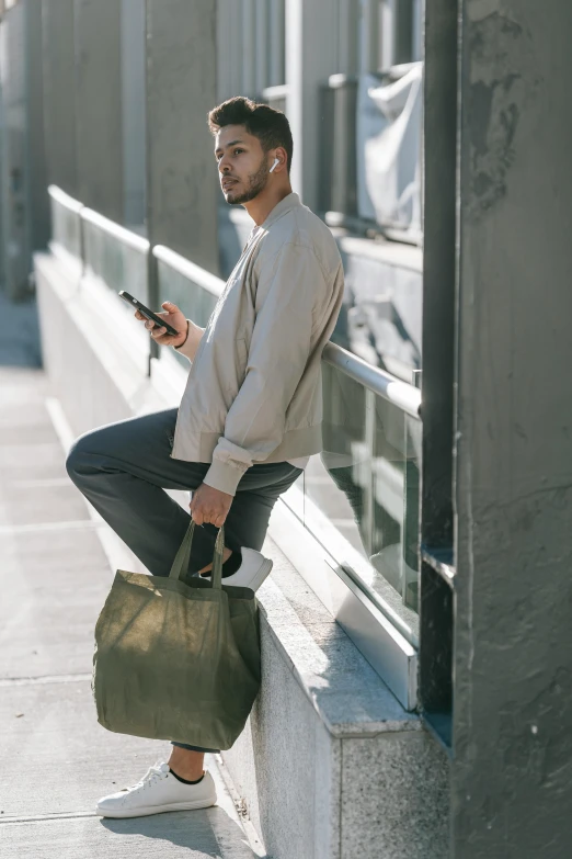 a man sitting on a ledge looking at his cell phone, by William Berra, trending on unsplash, renaissance, olive green slacks, holding a gold bag, wearing a baggy, standing in street