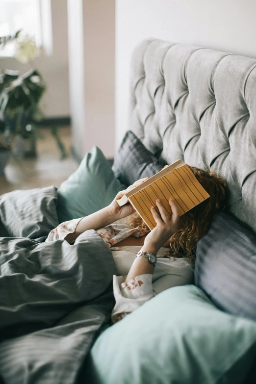 a woman laying in bed reading a book, with a wooden stuff, covered face, bedhead, sleep deprived