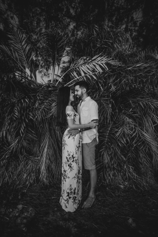 a couple standing next to each other in front of a tree, a black and white photo, by Matt Cavotta, pexels contest winner, chest covered with palm leaves, hugging her knees, pregnant, raphael personnaz