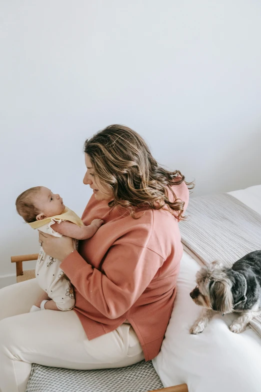 a woman sitting on a bed holding a baby and a dog, profile image, manuka, carefully crafted, cozy