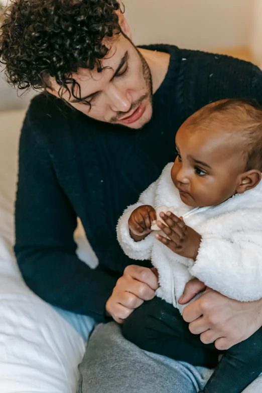 a man sitting on a bed holding a baby, inspired by Myles Birket Foster, pexels contest winner, mixed race, winter, curls, black