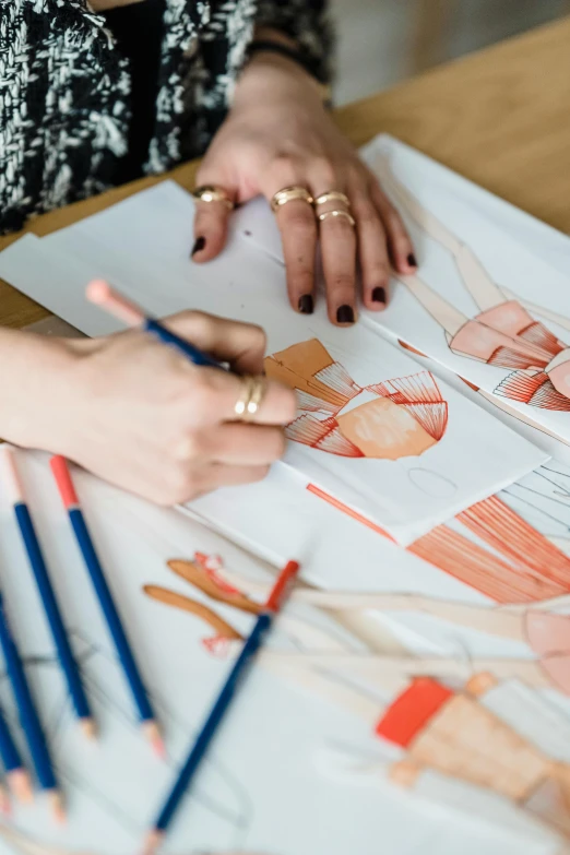 a woman sitting at a table drawing on a piece of paper, trending on pexels, process art, luxury fashion illustration, coloured in blueberra and orange, avant designer uniform, holding paintbrushes