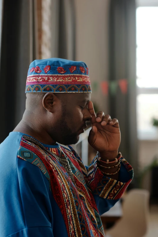 a man standing in a living room talking on a cell phone, an album cover, inspired by Ras Akyem, pexels contest winner, hurufiyya, red and blue garments, praying, wearing an elegant tribal outfit, african man