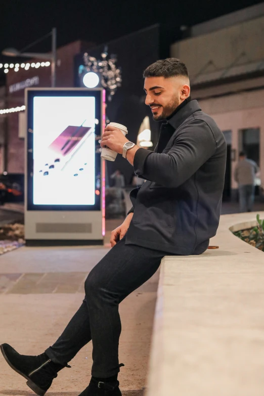 a man sitting on a ledge looking at his phone, trending on pexels, singer maluma, holding a drink, at a mall, zoomed out full body