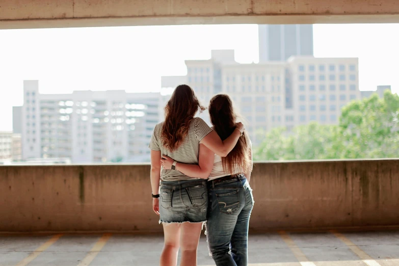 a couple of women standing next to each other, a picture, by Meredith Dillman, unsplash, set inside of parking garage, city in the distance, hugging, teenage girl