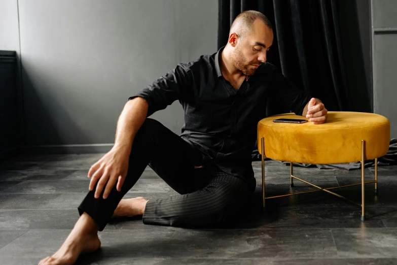 a man sitting on the floor in front of a yellow ottoman, inspired by Edi Rama, pexels contest winner, antipodeans, wearing a black shirt, performance, hasbulla magomedov, sleepy