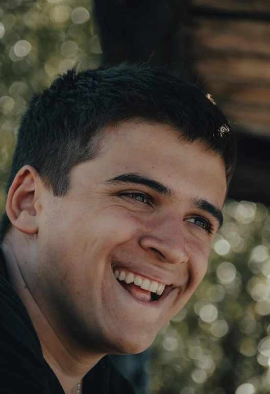 a close up of a person smiling with trees in the background, by Jacob Toorenvliet, young greek man, profile pic, large eyebrows, smol