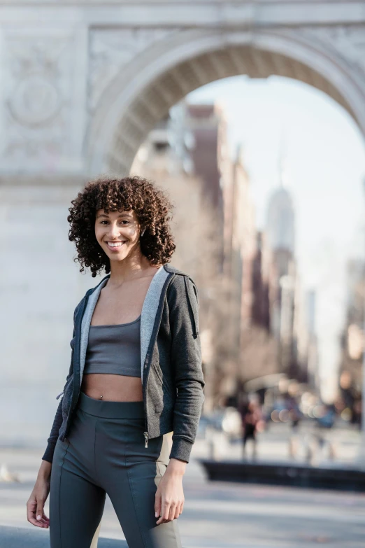 a woman standing in the middle of a street, curly bangs and ponytail, with abs, new york backdrop, smiling confidently