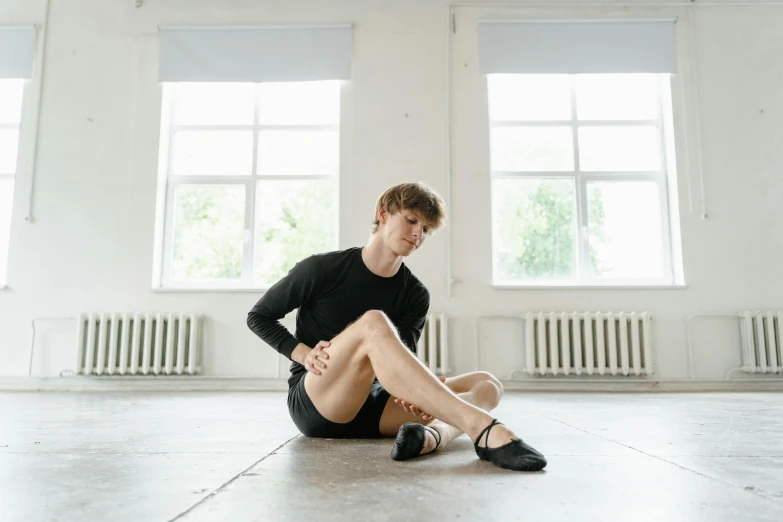 a man sitting on the floor with his legs crossed, by Elizabeth Polunin, trending on unsplash, wearing black shorts, dressed as a ballerina, alexey egorov, thin young male