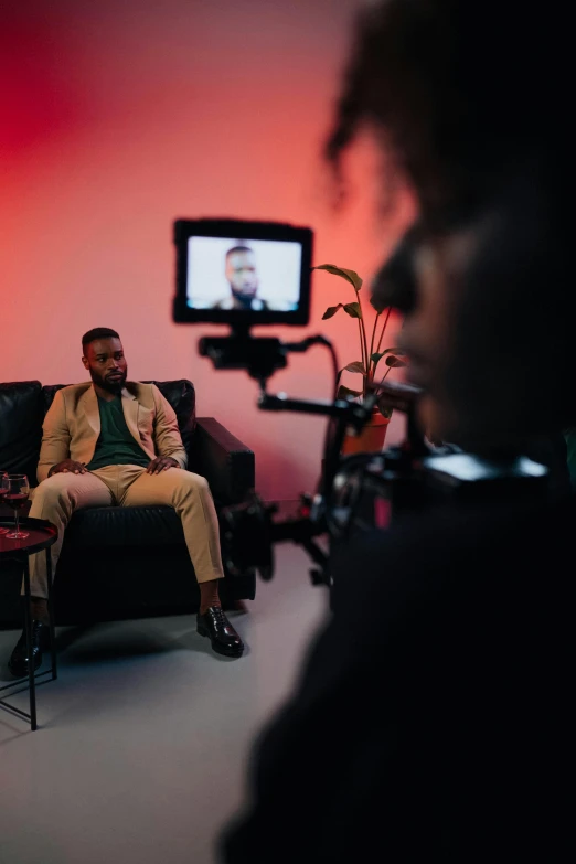 a man sitting on a couch in front of a tv, by Sebastian Spreng, pexels, giving an interview, man is with black skin, behind the scenes photo, ( ( theatrical ) )