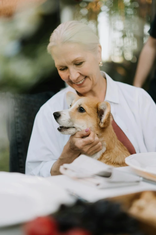 a woman sitting at a table with a dog, a portrait, by Nicolette Macnamara, unsplash, eating outside, jack russel dog, dinner is served, older woman