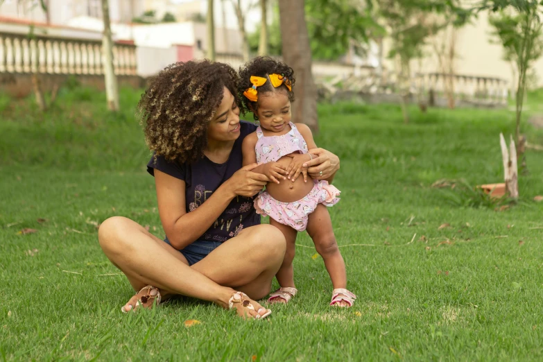 a woman holding a baby sitting on top of a lush green field, a portrait, by Samuel Silva, pexels, tan skin a tee shirt and shorts, avatar image, cute girls, with afro