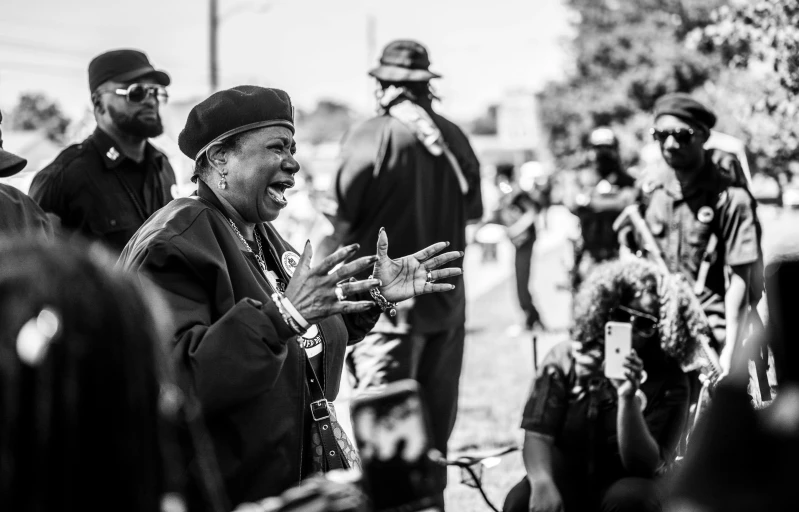 a man standing in front of a crowd of people, a black and white photo, by Dan Frazier, pexels, black arts movement, old lady screaming and laughing, gunfire, woman in black robes, memphis