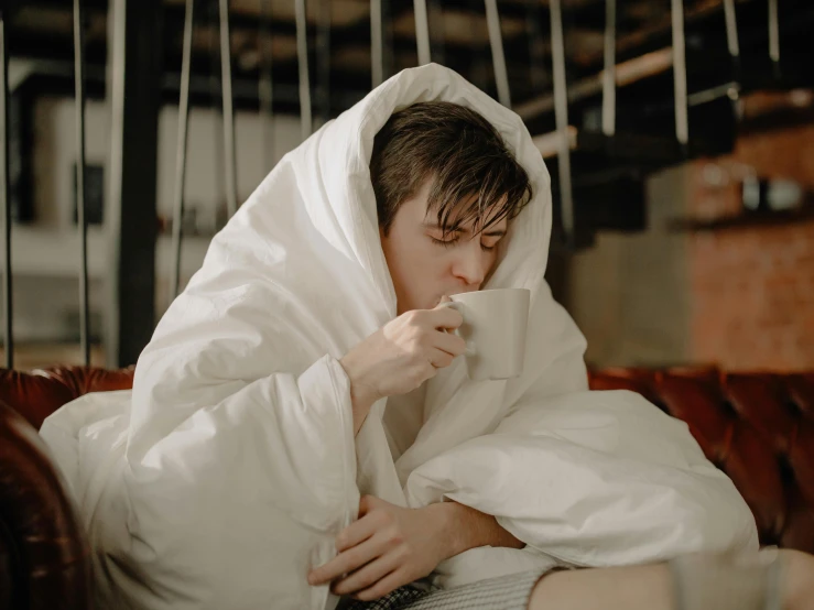 a woman sitting on a couch with a blanket over her head, pexels contest winner, happening, attractive man drinking coffee, declan mckenna, white bed, sweating profusely
