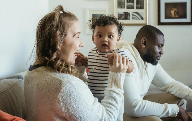 a man and woman sitting on a couch with a baby, pexels contest winner, incoherents, mixed race, looking her shoulder, brown, thumbnail