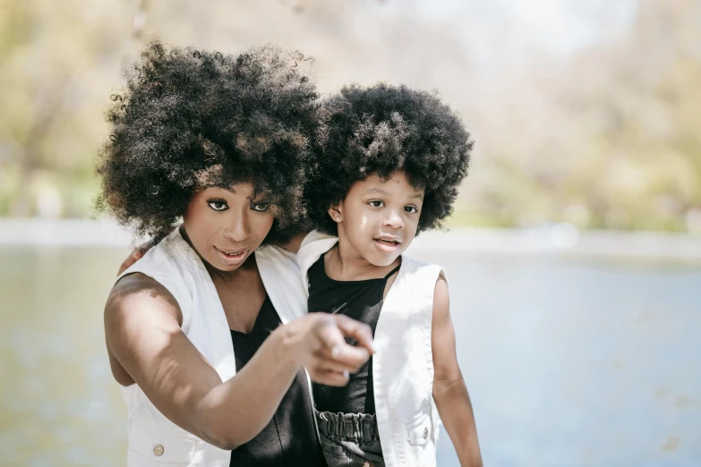 a woman and a child standing next to a body of water, pexels contest winner, afrofuturism, big hair, pointing at the camera, ivory and ebony, thumbnail