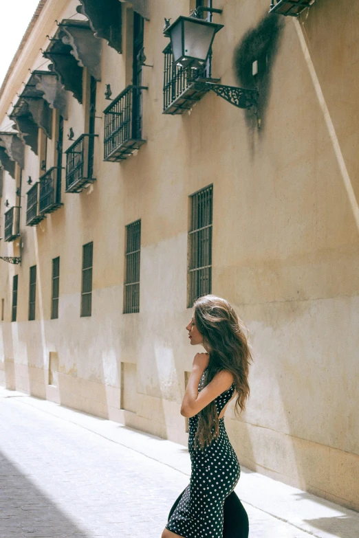 a woman walking down a street next to a tall building, inspired by Luis Paret y Alcazar, pexels contest winner, mid long hair, lady of elche, summer feeling, leaning on the wall