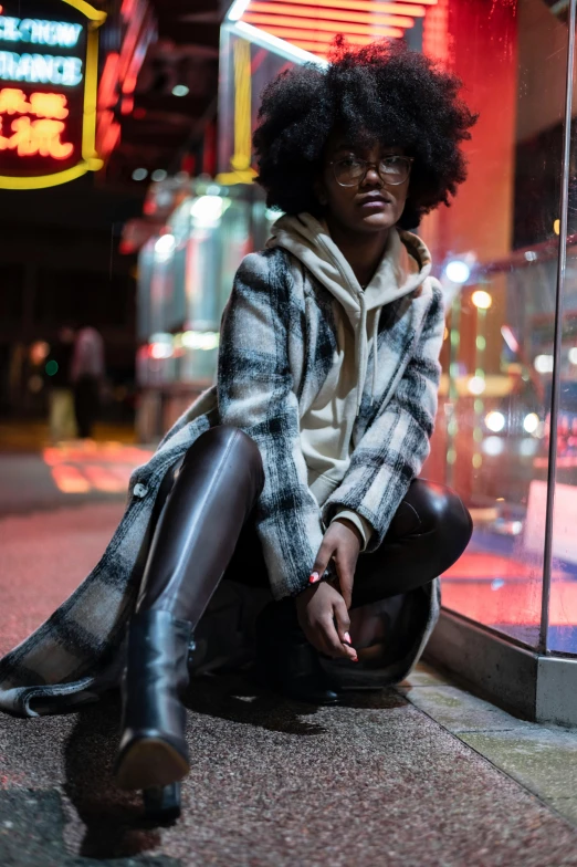 a woman sitting on the sidewalk in front of a store, by Jeffrey Smith, trending on pexels, wearing a long coat, tartan hoodie, black young woman, shot at night with studio lights