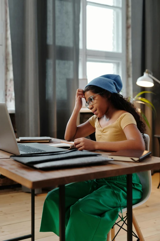 a woman sitting at a table with a laptop, with black beanie on head, hovering indecision, light skinned african young girl, gen z