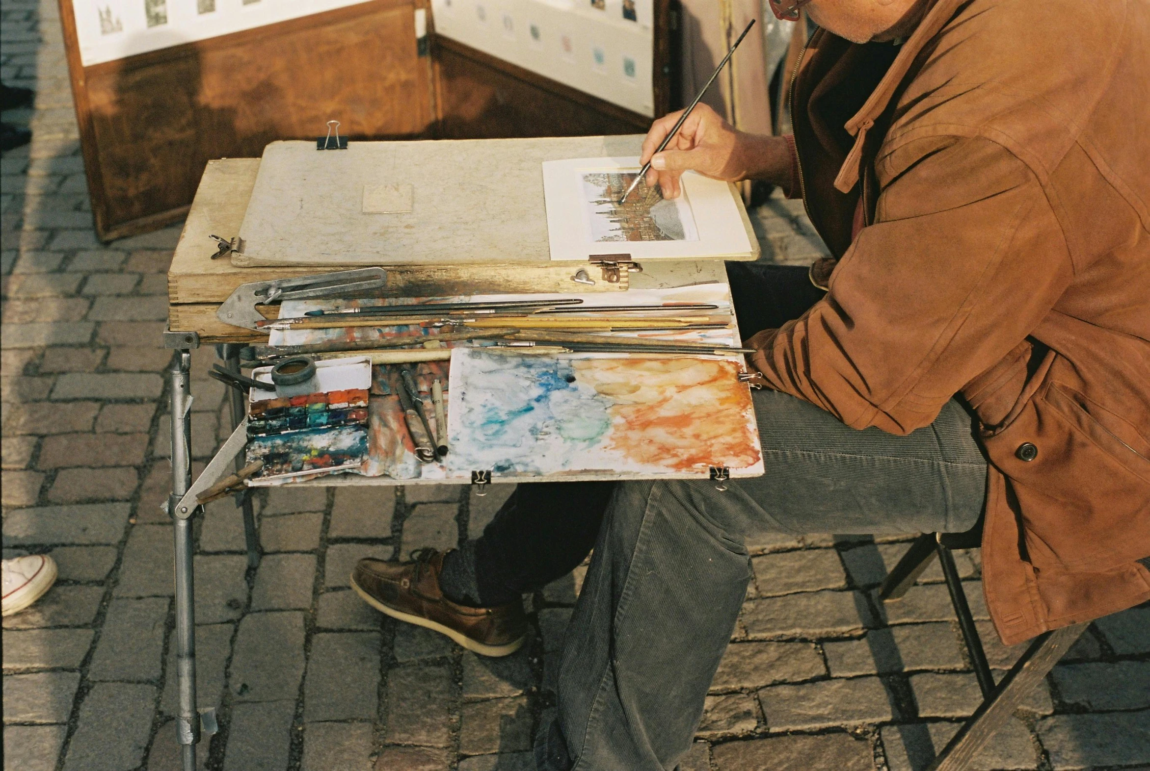 a man sitting at a table with a painting on it, pexels contest winner, paintbrush and palettes, street life, painted overalls, brown