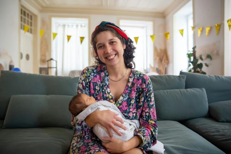 a woman sitting on a couch holding a baby, by Nina Hamnett, happening, relaxing and smiling at camera, simona sbaffi is the captain, h3h3, portrait image