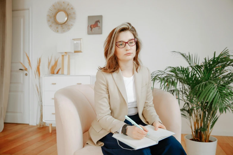 a woman sitting in a chair with a notebook and pen, a picture, trending on pexels, wearing a suit and glasses, beige, at the sitting couch, jovana rikalo