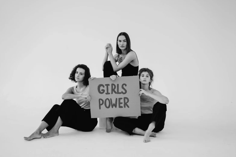 three women sitting next to each other holding a sign, an album cover, by Emma Andijewska, pexels, feminist art, power pose, powerful stance, portrait of gal gadot, no power