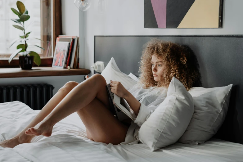 a woman sitting on a bed reading a book, by Carey Morris, pexels contest winner, curly haired, sexy :8, afternoon hangout, white bed