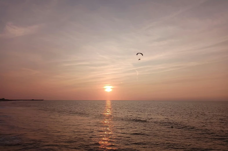 a person flying a kite over a body of water, on the beach during sunset, from the distance, evening time