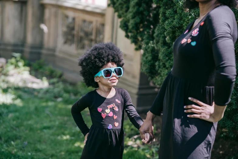 a woman standing next to a little girl wearing sunglasses, by Lily Delissa Joseph, pexels contest winner, funk art, black curly hair, black and cyan color scheme, she is wearing a black dress, schools