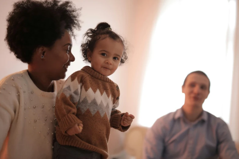 a woman holding a baby standing next to a man, pexels, light skinned african young girl, at home, 1 2 9 7, multicoloured